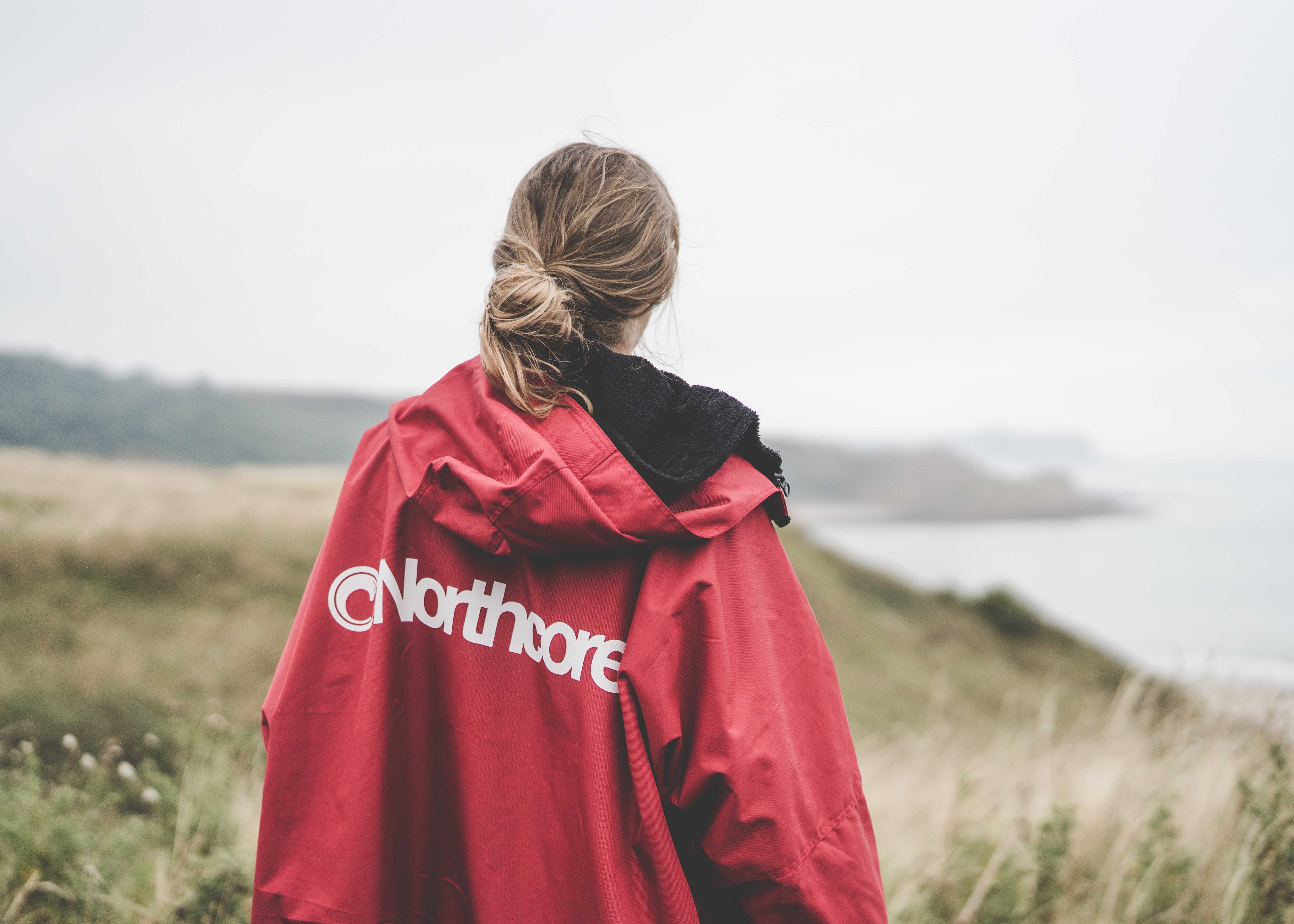 Woman seen from behind in a red Northcore change robe, walking through dunes above the sea